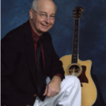 Larry Miller, sitting in front of a guitar with a blue background